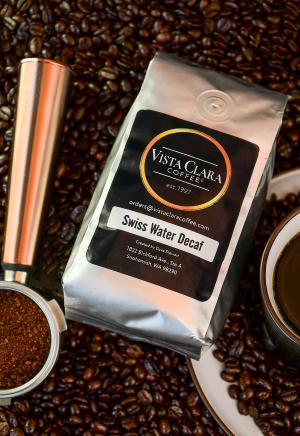 Vista Clara Coffee Swiss Water Decaf bag placed on coffee beans, accompanied by a portafilter filled with ground coffee and a cup of brewed coffee.