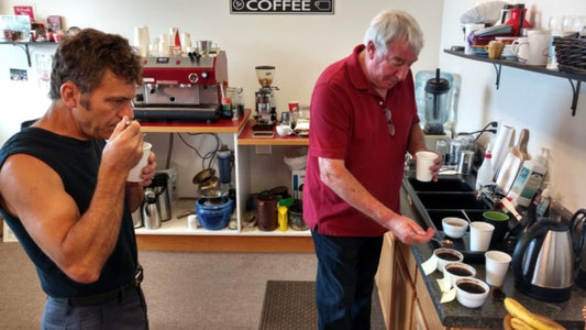 Man in black shirt and man in red shirt trying white cups of coffee in a kitchen with white walls.