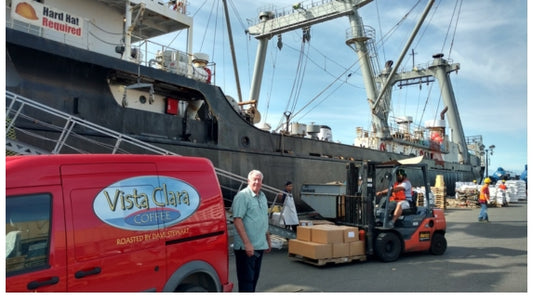 Man next to red van with yellow logo and light blue back. Large ship in background.