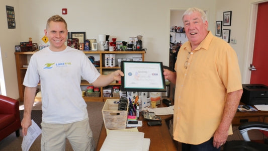 One man with white shirt and another man with yellow shirt holding white paper,  in tan background.